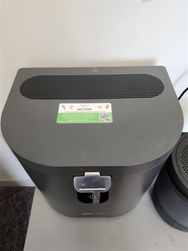Grey FoodCycler appliance with closed lid. Green and white "City of Nelson" sticker is adhered to the lid center of the appliance lid. 